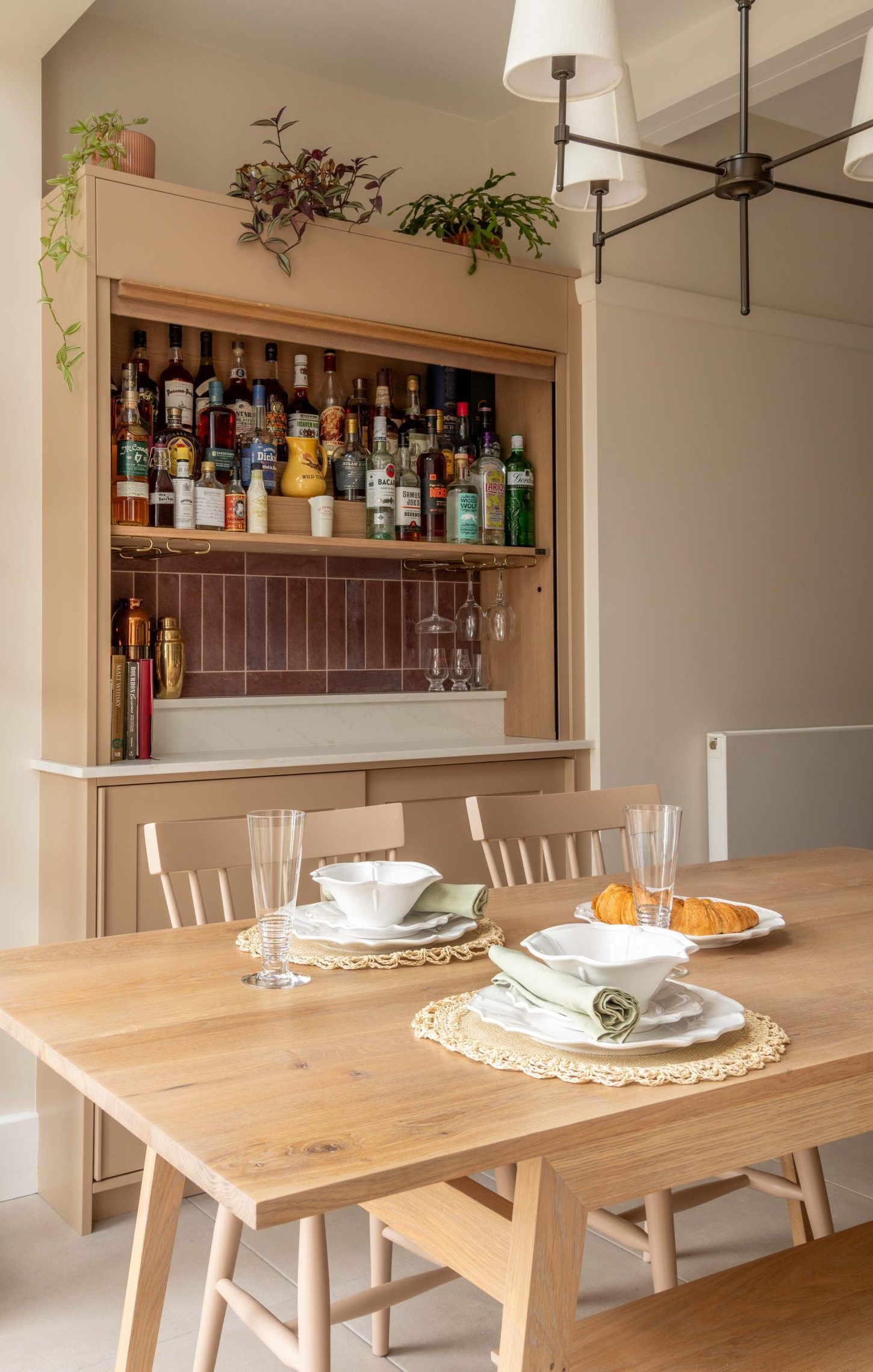 Bespoke concealed bar open, showing bottle shelving, dark tile splashback, brass glass rail and marble worktop beside oak dining table, designed by The House Ministry.