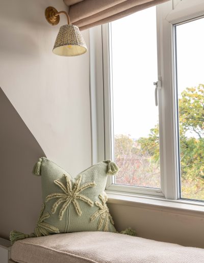 Bespoke window seat with open book storage, pull-out drawers, pink ceramic handles, brass wall light and garden view beyond, designed by The House Ministry.