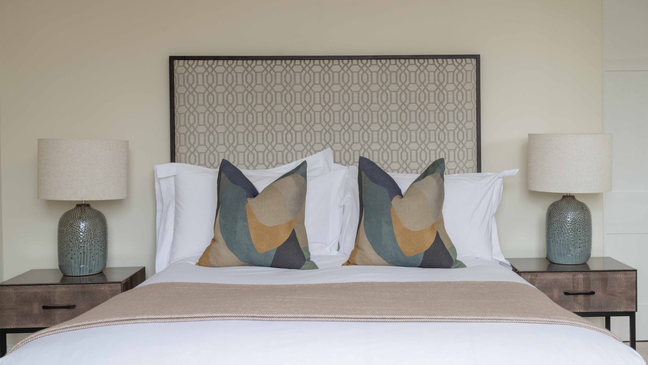 Bespoke Geometric Upholstered Headboard Detail | The House Ministry Bespoke geometric upholstered headboard with dark contrast piping, abstract cushions in teal and ochre, and textured ceramic bedside lamps, designed by The House Ministry.