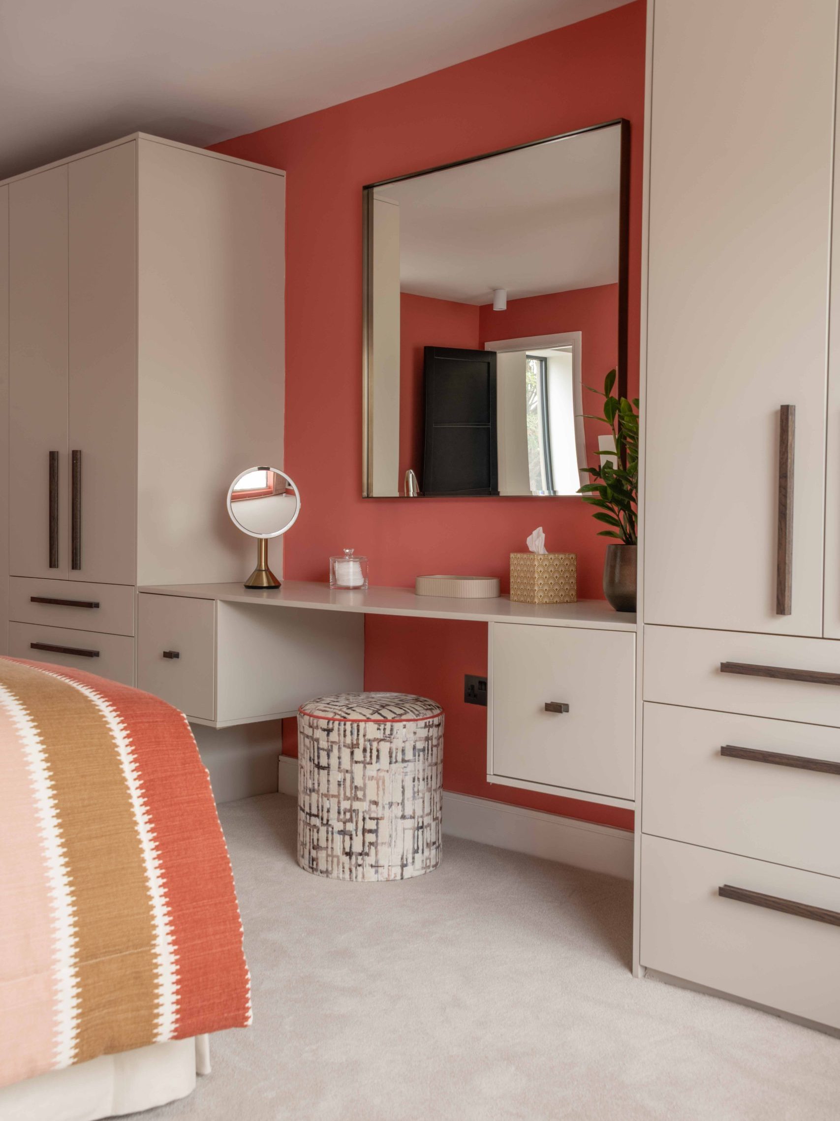 Fitted Dressing Area | The House Ministry Fitted dressing area with coral accent wall, built-in desk, round vanity mirror and patterned stool between fitted wardrobes
