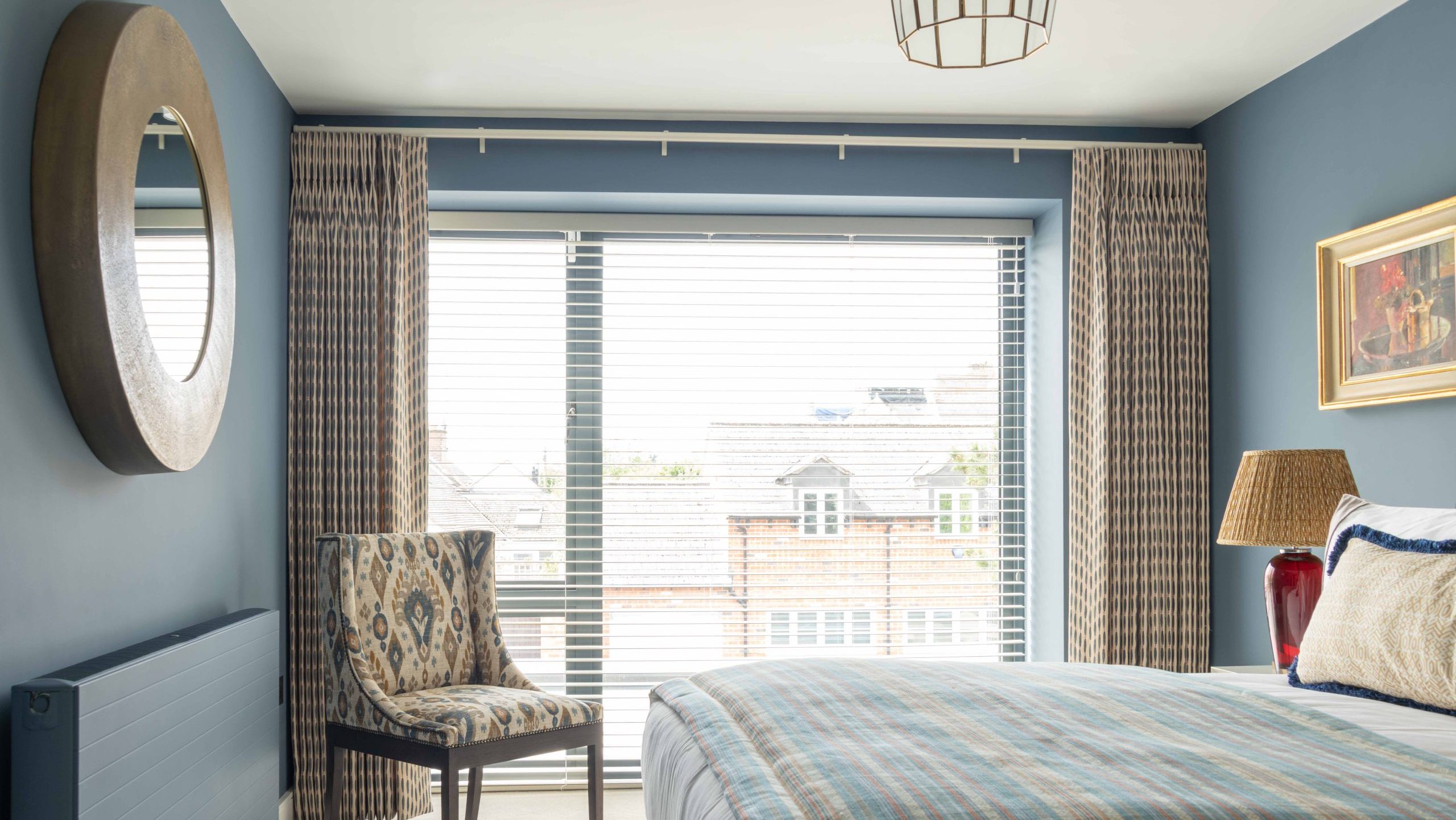 Guest Bedroom Full View | The House Ministry Guest bedroom with round bronze mirror, ikat-patterned chair, patterned curtains and a striped bedspread