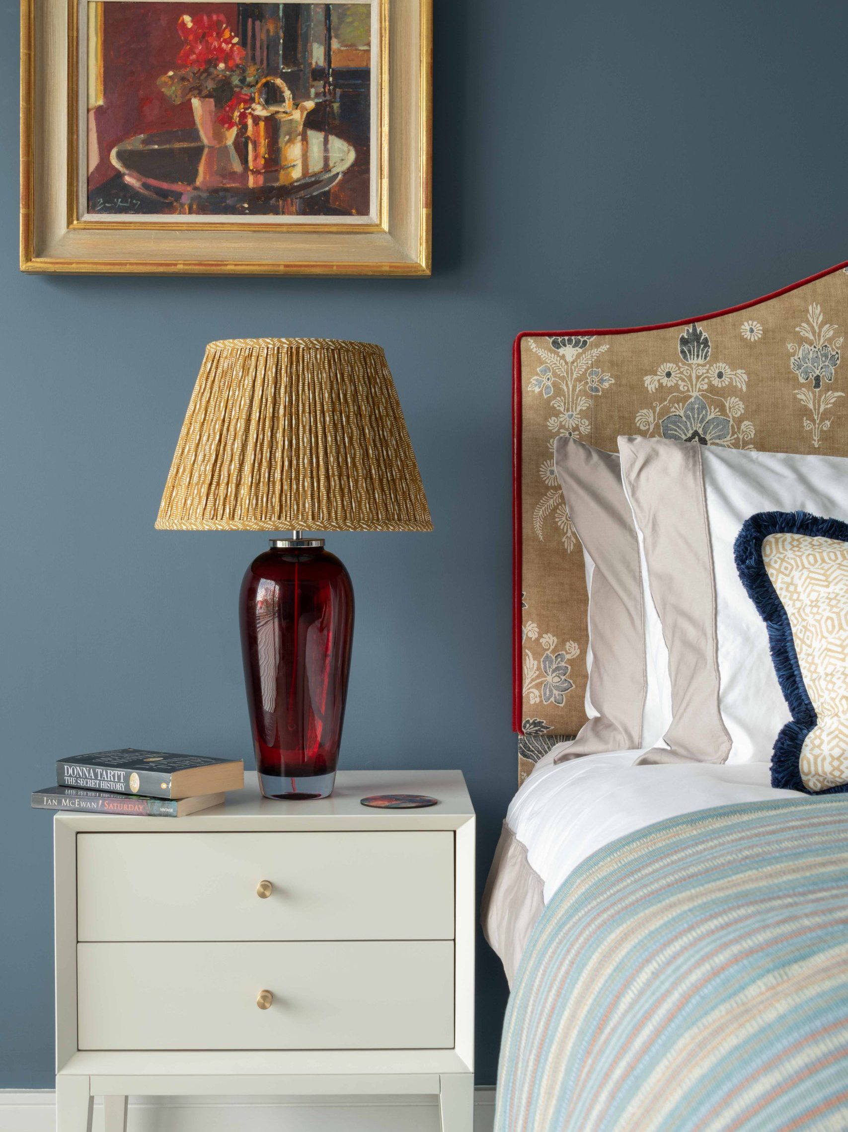 Bedside Styling Detail | The House Ministry Bedside table with a red glass lamp, pleated patterned lampshade, gilt-framed artwork and stacked books