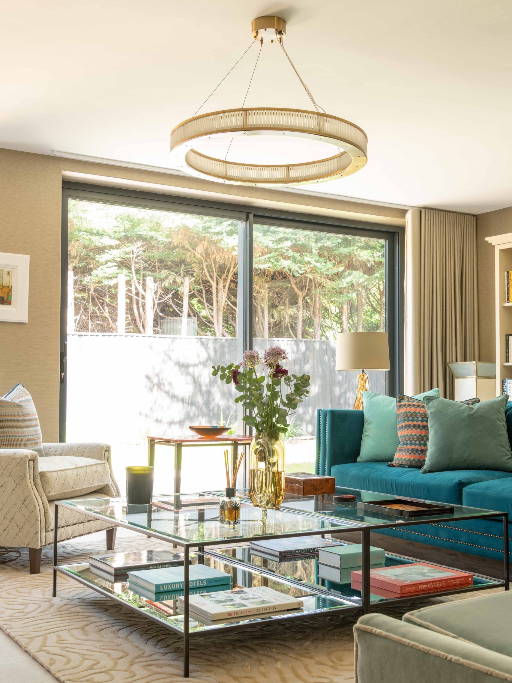 Living Room with Statement Pendant Light | The House Ministry Living room with a ring-style brass and glass rod pendant light above a glass coffee table, teal sofa and sliding garden doors
